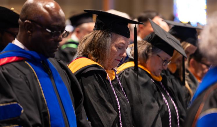 20240909_convocation_0006 A group of faculty members, dressed in black academic regalia with colorful hoods, bow their heads in prayer or reflection during ETBU's fall 2024 Convocation chapel