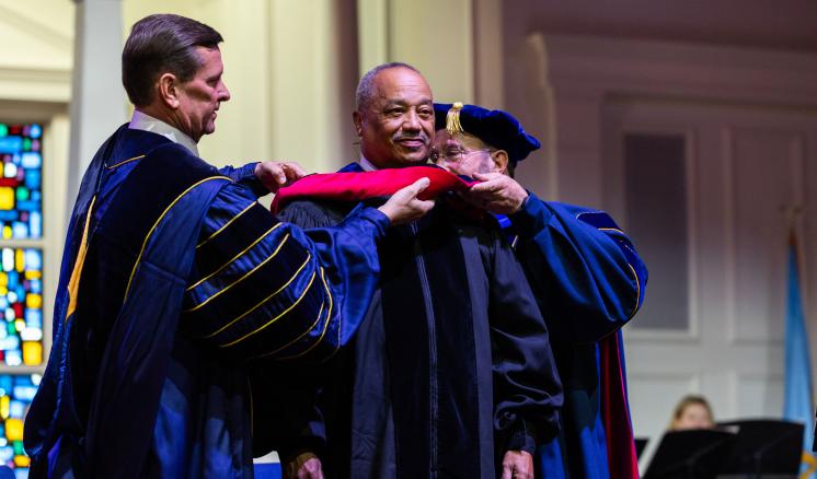 20240909_convocation_0314 Two faculty members, wearing academic regalia with doctoral hoods, are seen placing a bright red hood over the shoulders of an honoree