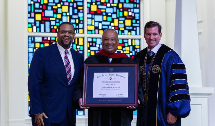 20240909_convocation_0444 The honoree, holding a framed honorary degree, stands between two university officials, all smiling. They are standing on stage in front of a brightly lit stained glass window, with the university officials dressed in dark blue and black regalia. The plaque reads, "East Texas Baptist University" at the top.