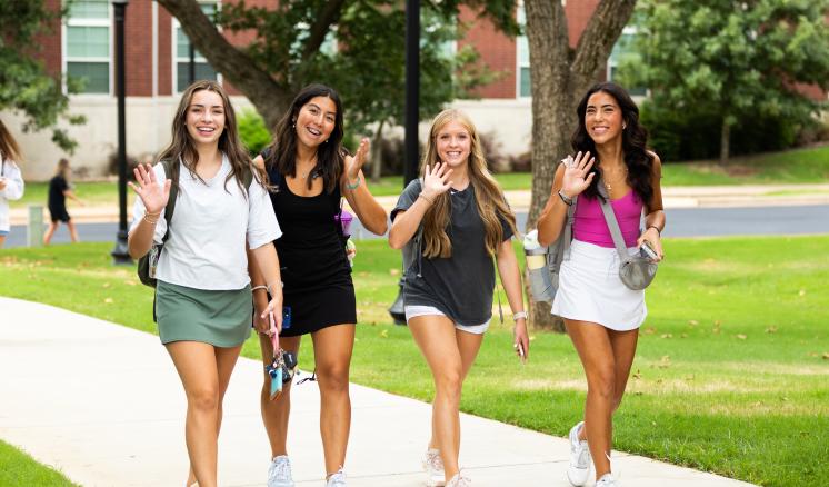 Four young women walking on a college campus, smiling and waving towards the camera. They are dressed in casual attire and carrying backpacks. The background shows green lawns, trees, and part of a brick building.
