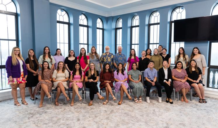 Polly Cargill Scholarship Reception 2024 Group photo of a diverse team of individuals at East Texas Baptist University, posed in a bright, spacious room with large arched windows. The group consists of men and women, dressed in a variety of professional and semi-casual attire, seated and standing in two rows.
