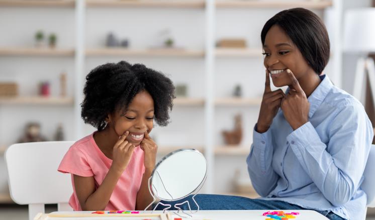 A child and speech-language pathologist sit together at a table, both smiling warmly and pointing to their cheeks in front of a small mirror.