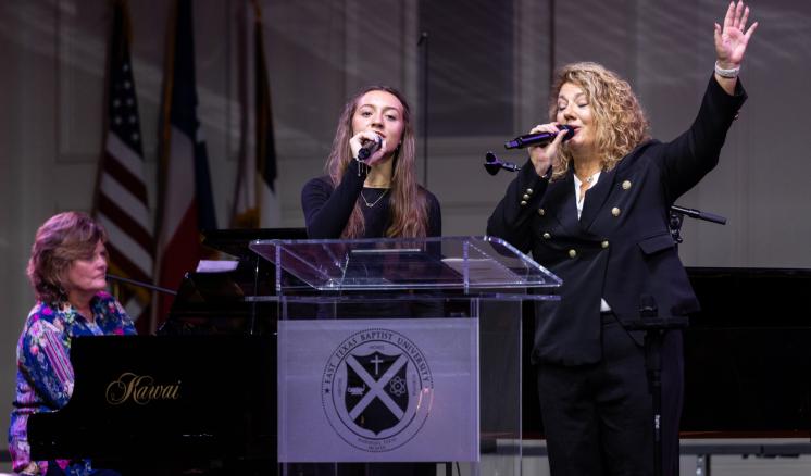 Three women performing on stage, one playing a piano, and two singing behind a podium with several flags in the background.