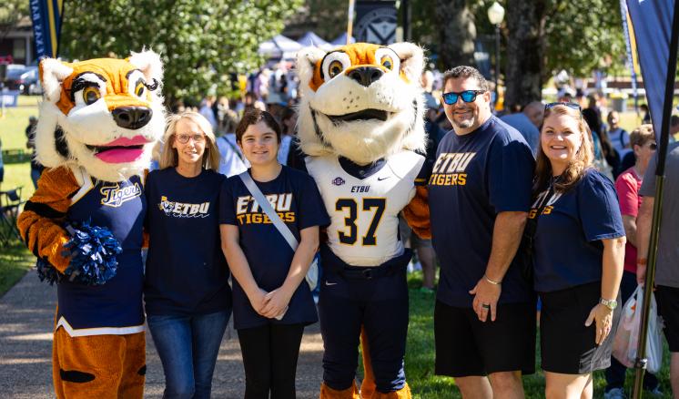 A group of people, including two university mascot tigers, pose together at an outdoor campus event, all wearing school spirit shirts.