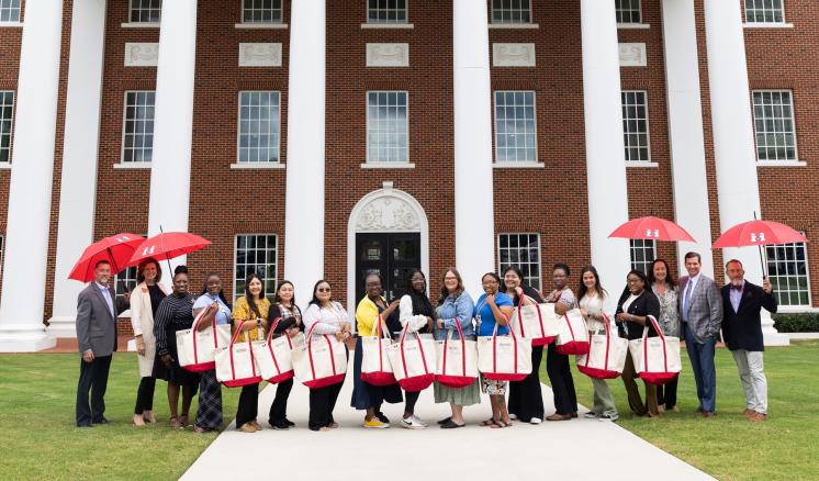 img_1 Group of people holding tote bags and red umbrellas, standing in front of a large brick building with white columns and windows.