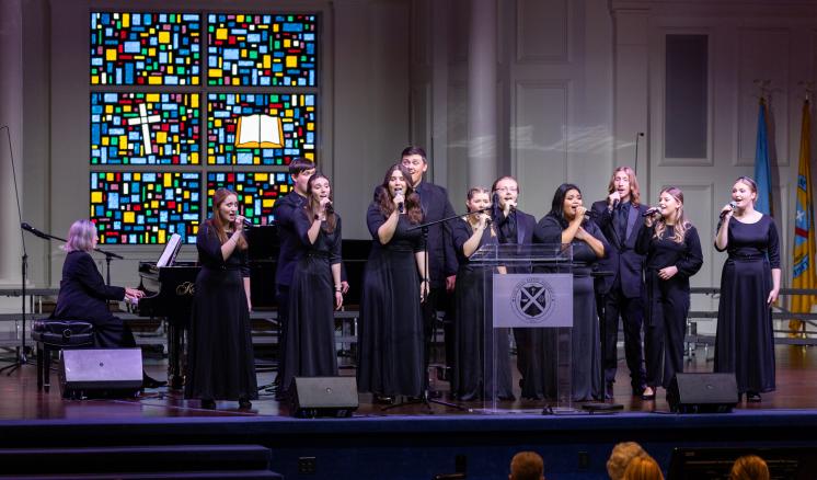  A group of eleven people in black formal attire perform on a stage with a pianist, under a colorful stained glass window.