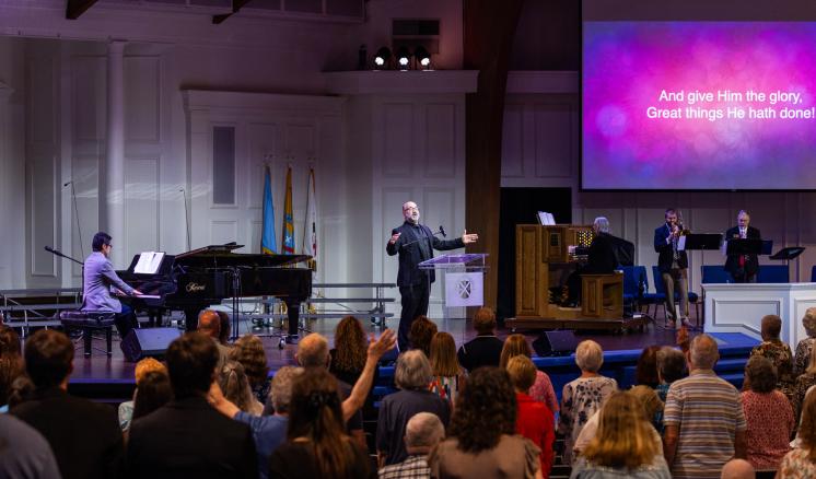  A stage with people playing piano, speaking, and addressing an audience in a church or auditorium setting.