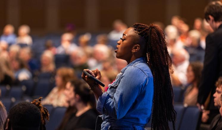 img_4 A person stands and speaks into a microphone in a crowded indoor venue with rows of seating.