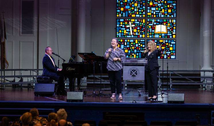 Three people performing on a stage with a piano and a colorful stained glass window in the background.