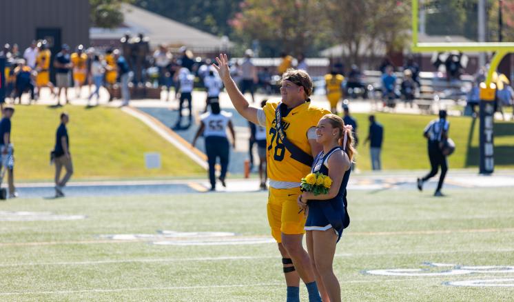 A football player and a cheerleader stand together on the field during a homecoming ceremony, with the player pointing towards the stands.
