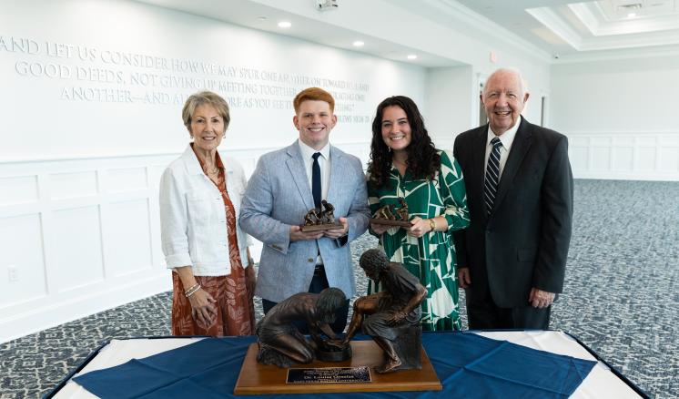 Riley Servant Leader Award 2024 A group of four people stands smiling in front of a table displaying a bronze statue of Jesus washing a disciple's feet. The group consists of two men and two women, all dressed formally.