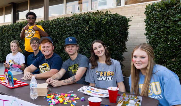 A group of young adults at a table with craft supplies, outdoors by a hedge and building.