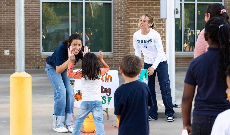 Two volunteers engage with children at a fall festival activity, sharing high-fives in front of a sign that reads "Pumpkin Bag Toss."