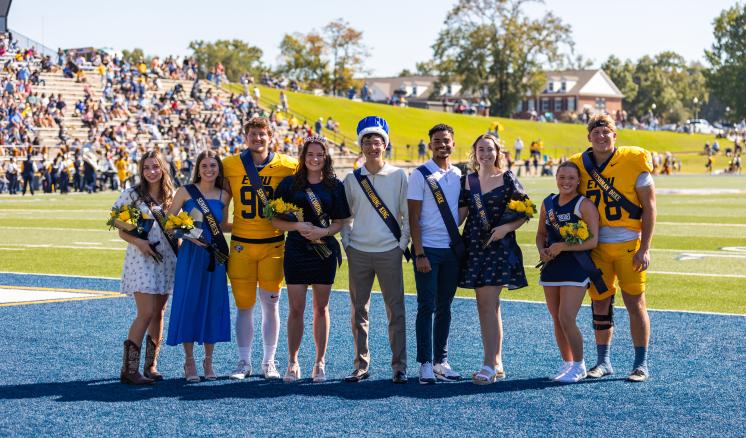 A group of students, including football players and homecoming royalty, poses together on a football field during a sunny homecoming event.