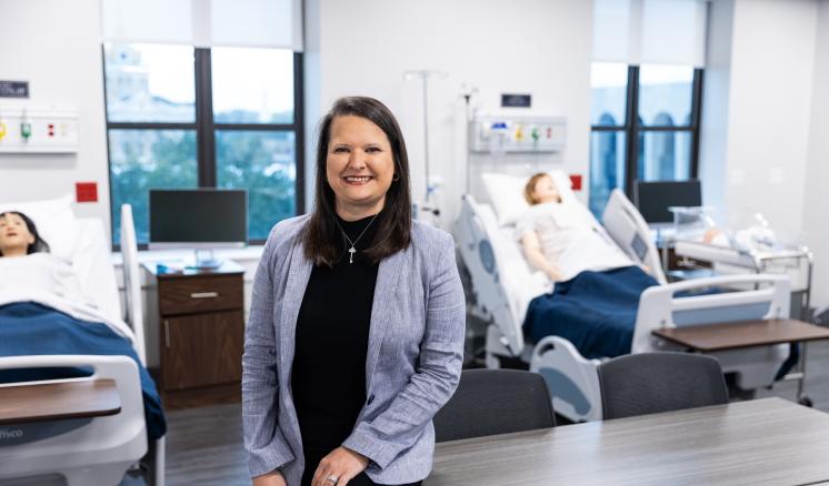 Smiling woman in a professional outfit stands in a modern nursing simulation lab, with hospital beds, mannequins, and medical equipment visible in the background.
