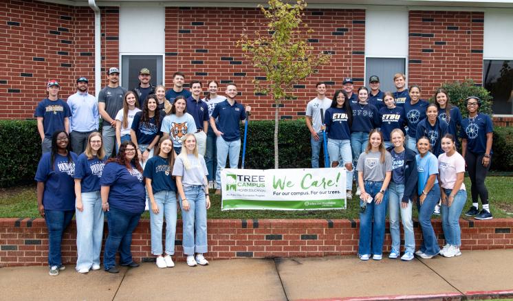 Students stand around a freshly planted tree with a banner between them in honor of Texas Arbor Day