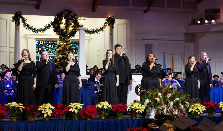 Singers in black attire perform on a festive stage with poinsettias and a decorated wreath.