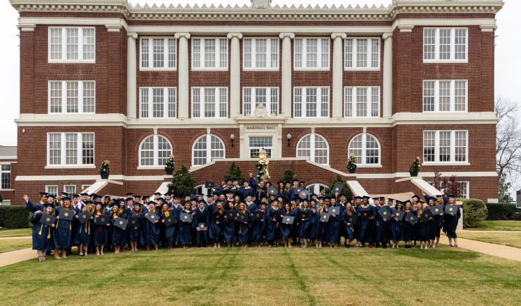 A large group of graduates in blue robes poses in front of a historic red-brick university building.
