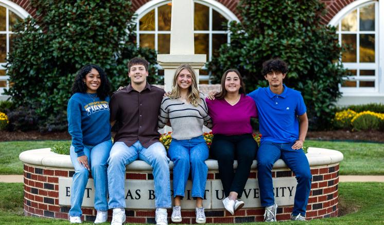 Spring 2025 Enrollment Five college students sit on a circular raised flower bed surrounding ETBU's Light on the Hill