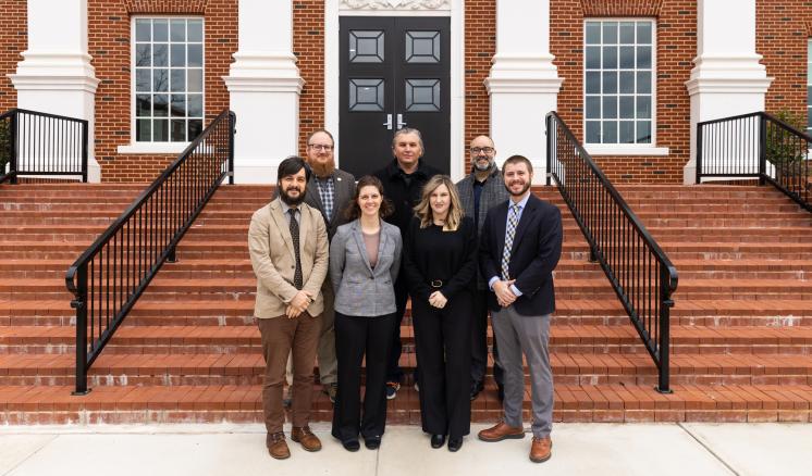 A group of seven formally dressed individuals standing on steps in front of a brick building with double black doors.