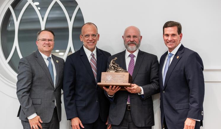 Four men in suits stand together, one holding a statue with a wooden base.