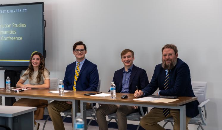 Four people sit behind a table in professional attire, a TV screen with a presentation is seen in the background