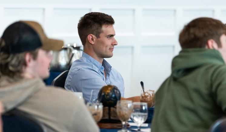 3 men are seated assumably listening to a speaker at a table with a blue tablecloth with a small globe as a centerpiece