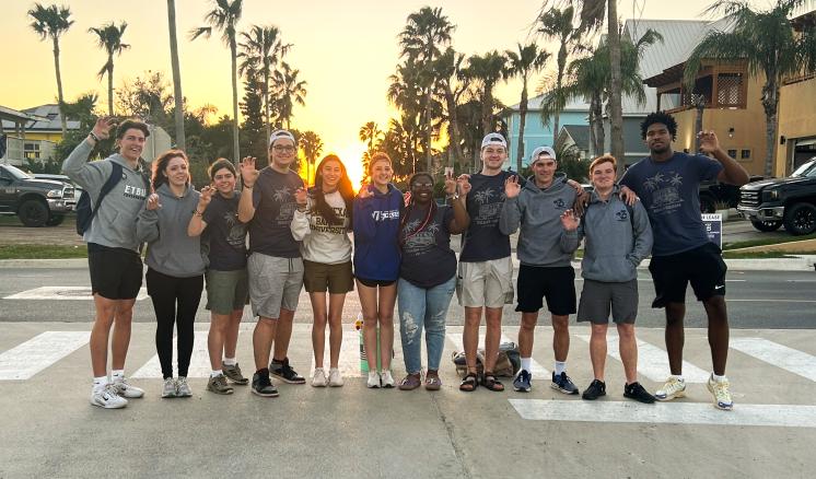 A group of people stood outside near a street with palm trees and a sunset behind them