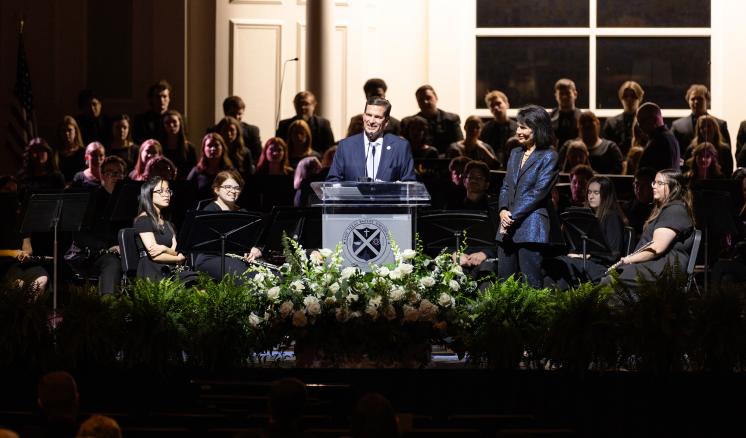 Many people on a stage in a dark lit room with a light fixed on a speaker stood at a podium