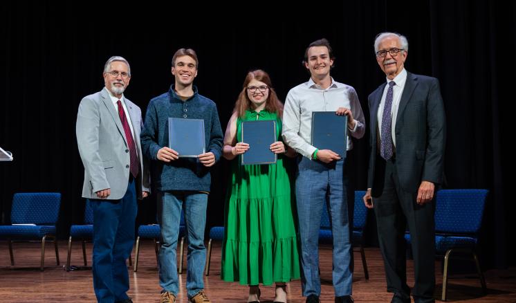 5 people stood on a dimly lit stage with blue chairs behind them, holding awards