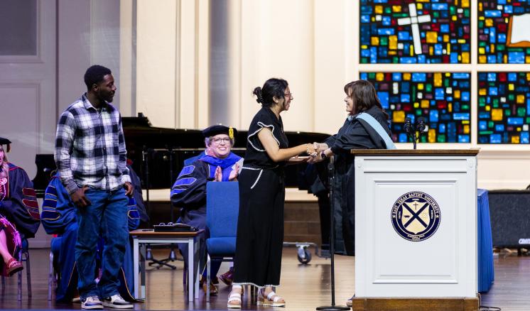 A woman receiving an award from another woman dressed in academic regalia stood behind a podium on a well-lit stage