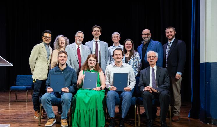 A group of people sitting and standing on a dimly lit stage smiling for a group photo