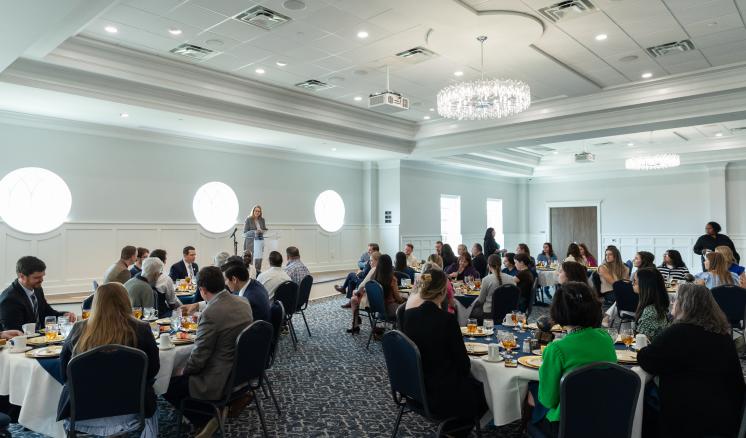 A woman stood in front of a podium speaking to a well lit room of people eating food