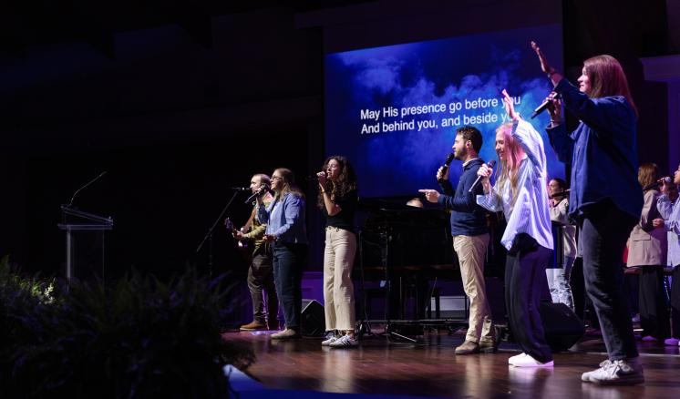 a group of people stood on a stage singing in front of a project screen. The whole scene is dimly lit