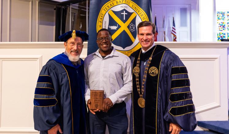 3 people stood in front of the seal for East Texas Baptist University, in a well-lit room wearing academic regalia