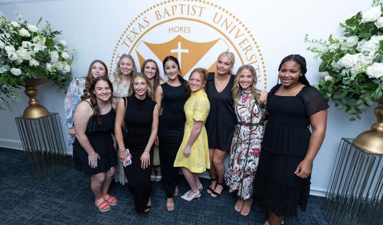 a group of people standing in a well-lit room with the seal of East Texas Baptist University behind them