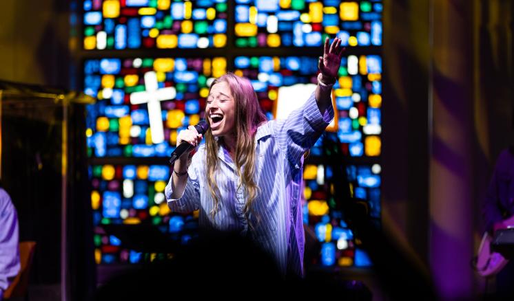A woman stood smiling and laughing in front of a stained glass window with a cross etched into it