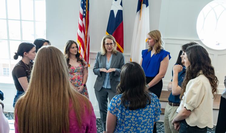A group of people stood in a well lit room in front of flags talking to each other in a circle
