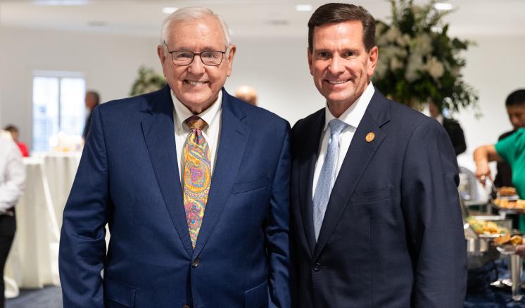 2 men stood together smiling inb a brightly lit room with flower centerpieces behind them