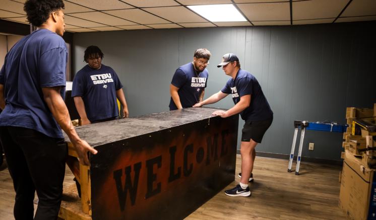 4 people carrying a large wooden welcome sign