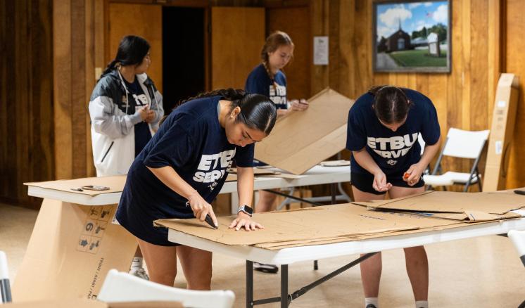 4 people stood over a table cutting cardboard on plastic camp tables