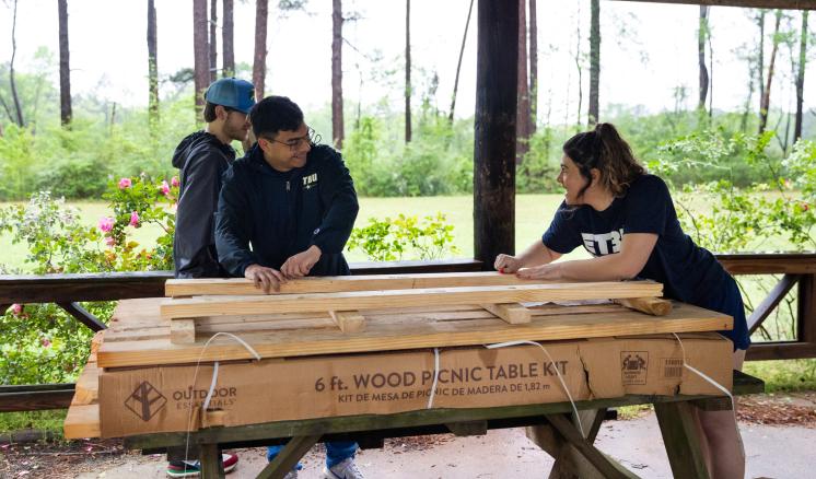 2 people stood in front of a screen window unpacking a wooden picnic table box