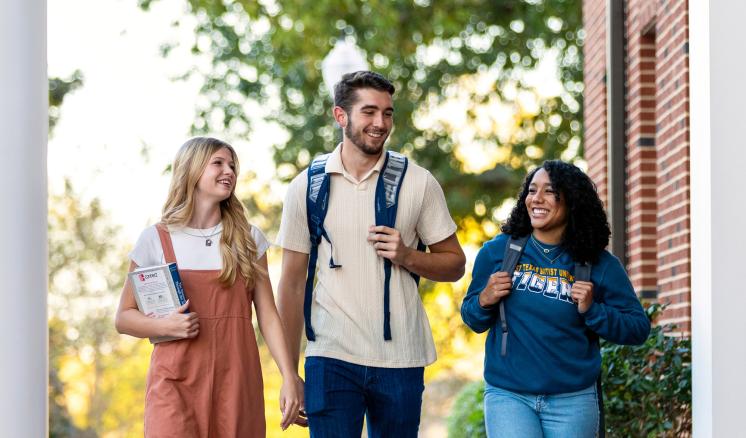 Three college students walk and talk together on campus, smiling and carrying backpacks and books.