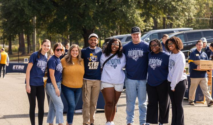 A group of smiling young adults and one older adult pose together outdoors in ETBU apparel during a campus event.