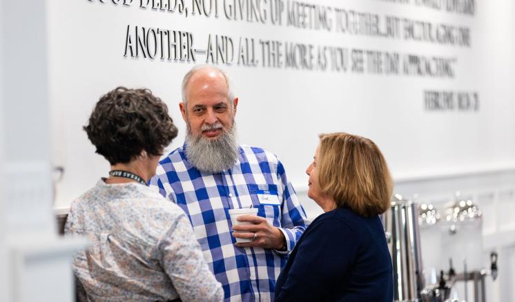 A man and 2 women looking at eachother standing in a circle in front of a board with a bible verse