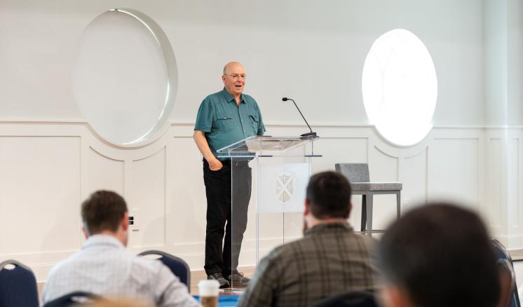 A speaker stood at a podium in a brightly lit room while a crowd of onlookers listen