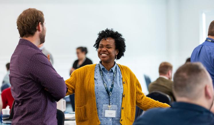 A lady stood talking to a man and smiling in a bright room full of people