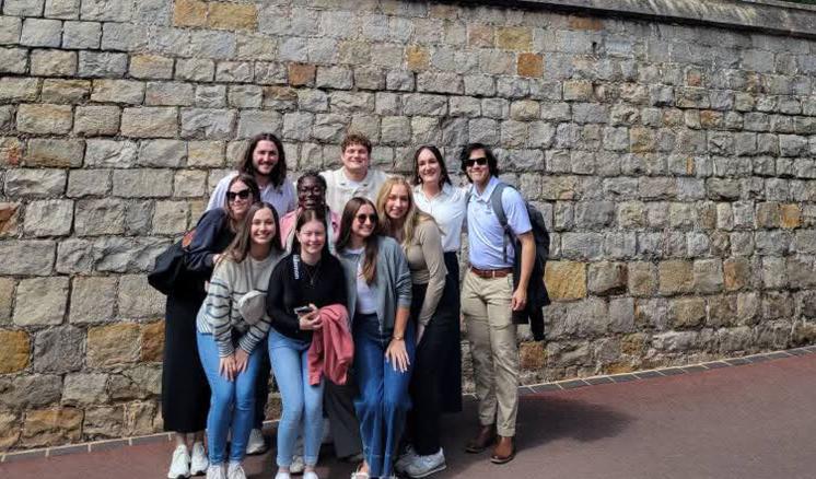 A group of people standing outside in daylight beside a brick wall with a large brick building in the background