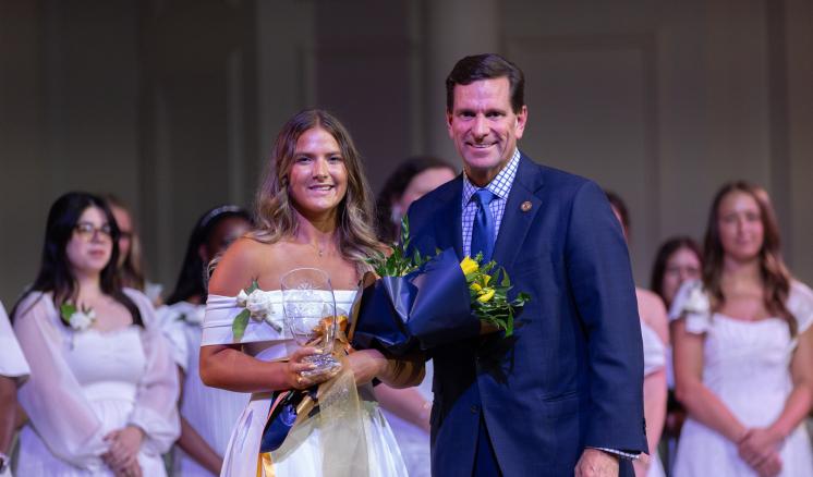 2 people in formal clothing smiling in front of a large group of people in white clothing behind them