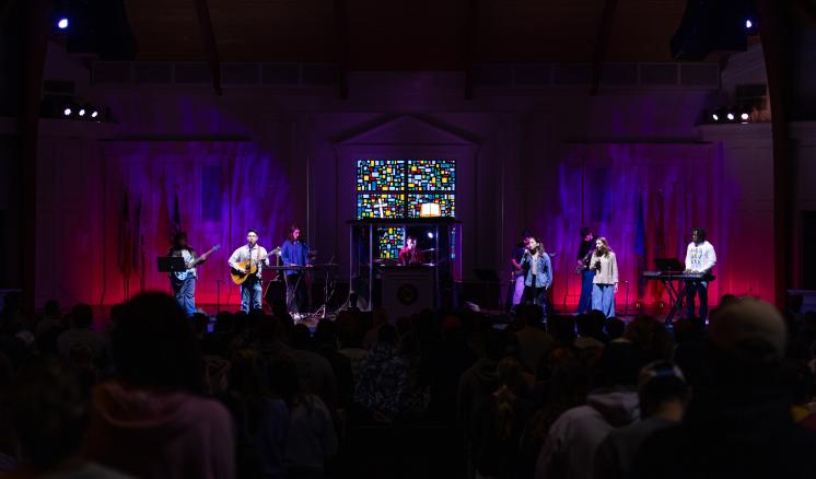 6 people standing on a dimly lit stage holding instruments in front of a large auditorium crowd